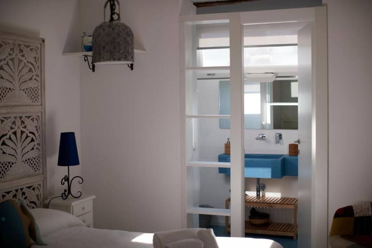 A bedroom with a bed, a nightstand, a lamp, a wardrobe with a mirror, and a blue bathroom sink.