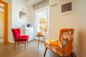 Una habitación con una silla roja, un sillón estampado, una mesita con una taza y una ventana con vistas a la vegetación.