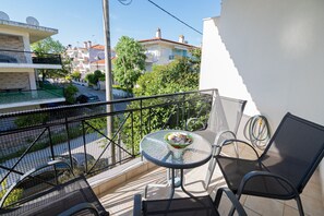 En balkon med et bord og stole, en skål med frugt, og udsigt til bygninger og træer.