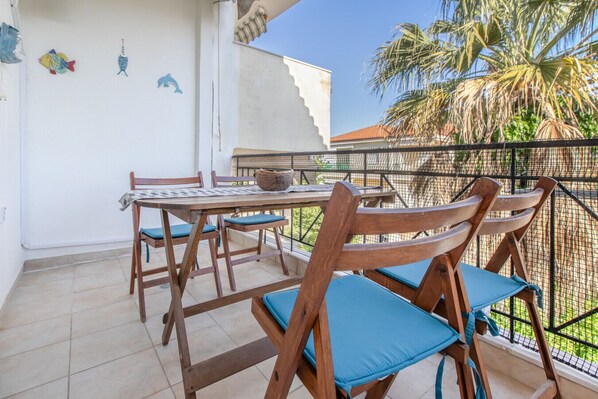 En balkon med et bord og stole, udsigt til palmer og dekorativ fiskekunst på væggen.
