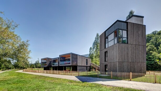 Een modern gebouw met twee verdiepingen, een plat dak, grote ramen en een houten omheining rond het terrein.