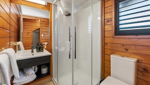 Une salle de bain aux murs lambrissés, dotée d’une douche en verre, de toilettes et d’un lavabo avec des articles de toilette.
