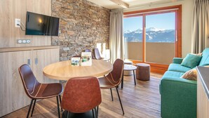 A modern room with a stone wall, wooden ceiling, a dining table with chairs, a sofa, and a large window with a mountain view. - Group Mountain Stay for Six in Doucy (La Léchère)