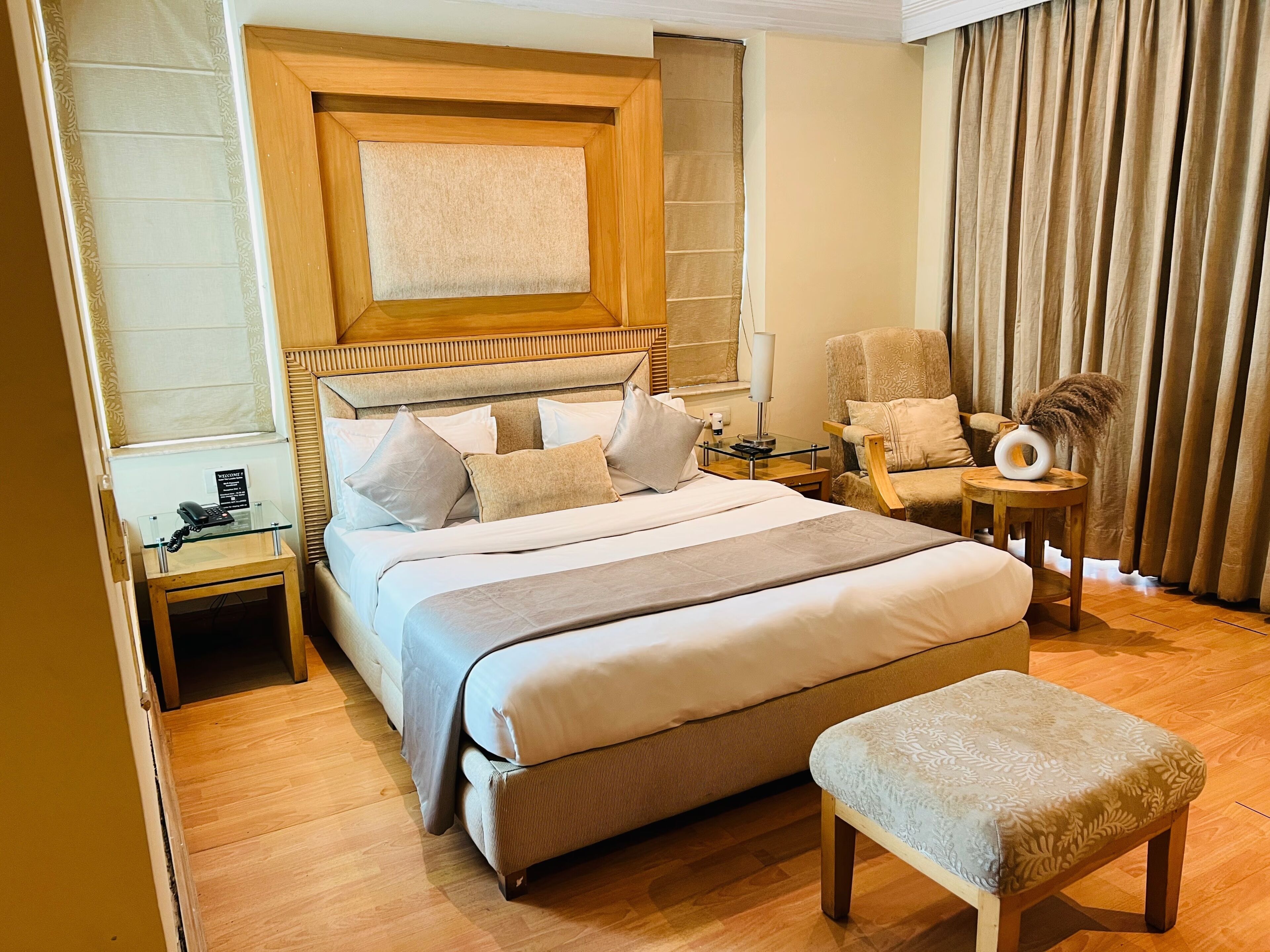 A hotel room with a large bed, a wooden headboard, a bedside table with a lamp, a chair, a small round table with a vase, and a telephone on a glass table.