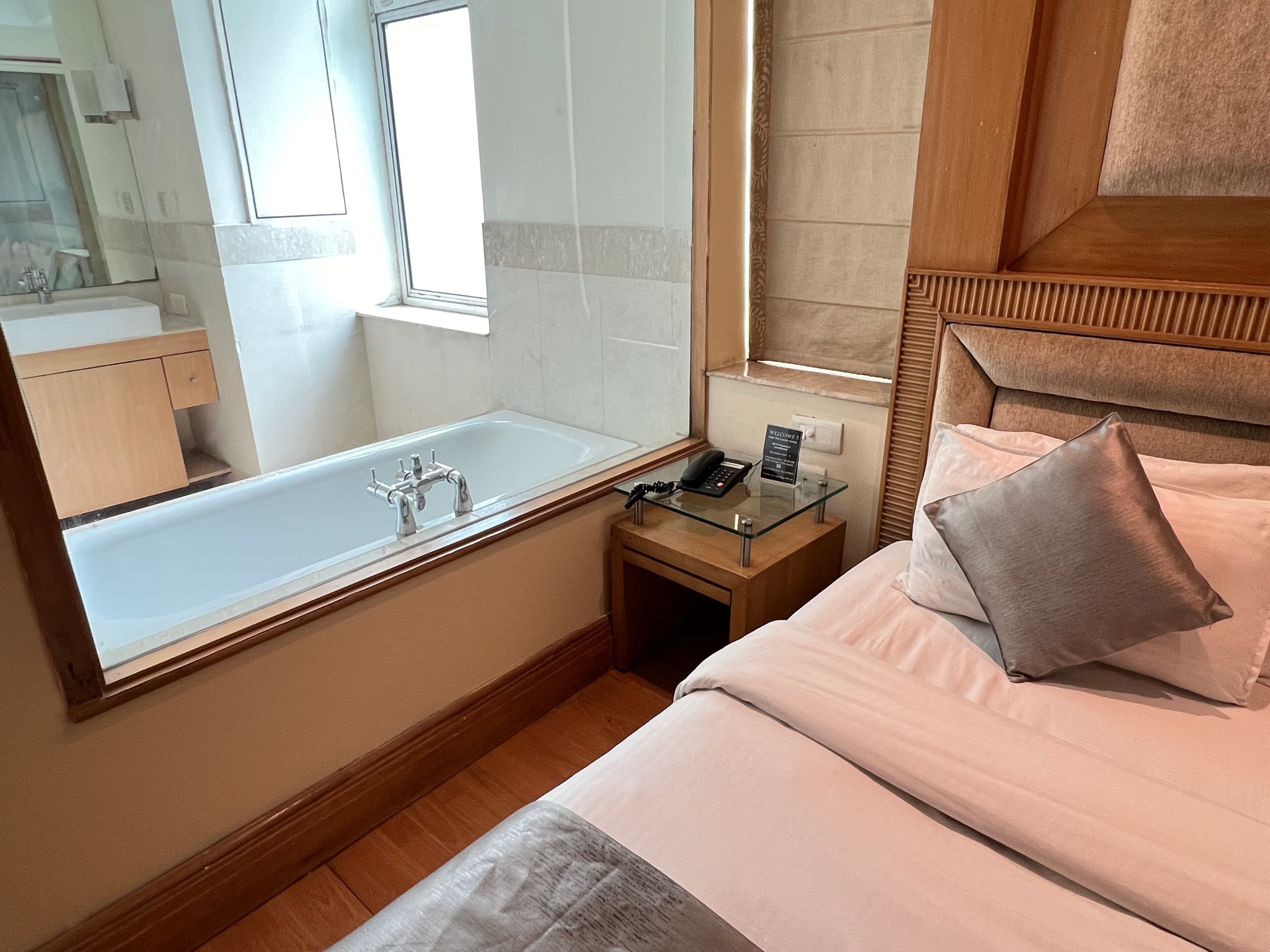 A hotel room with a large bathtub, a bedside table with a phone and a sign, a bed with pillows, and a window.