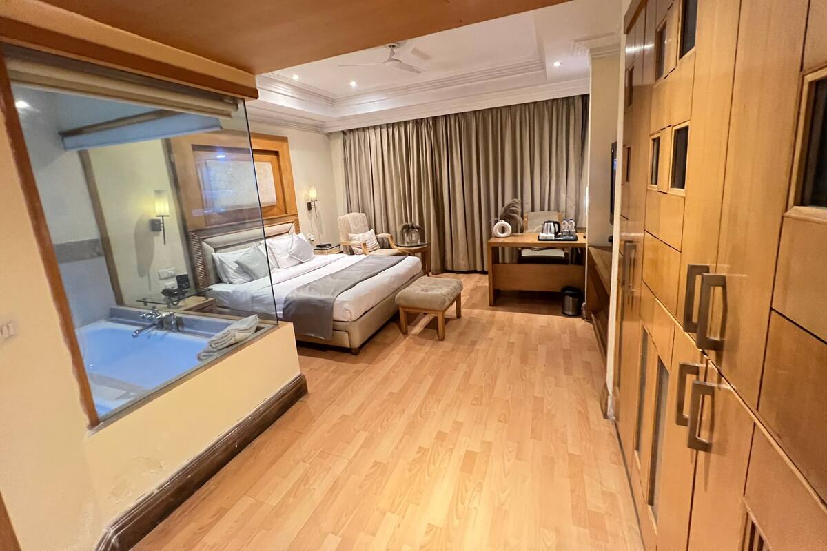 A hotel room with a bed, desk, chair, and a view of the city through a large window.