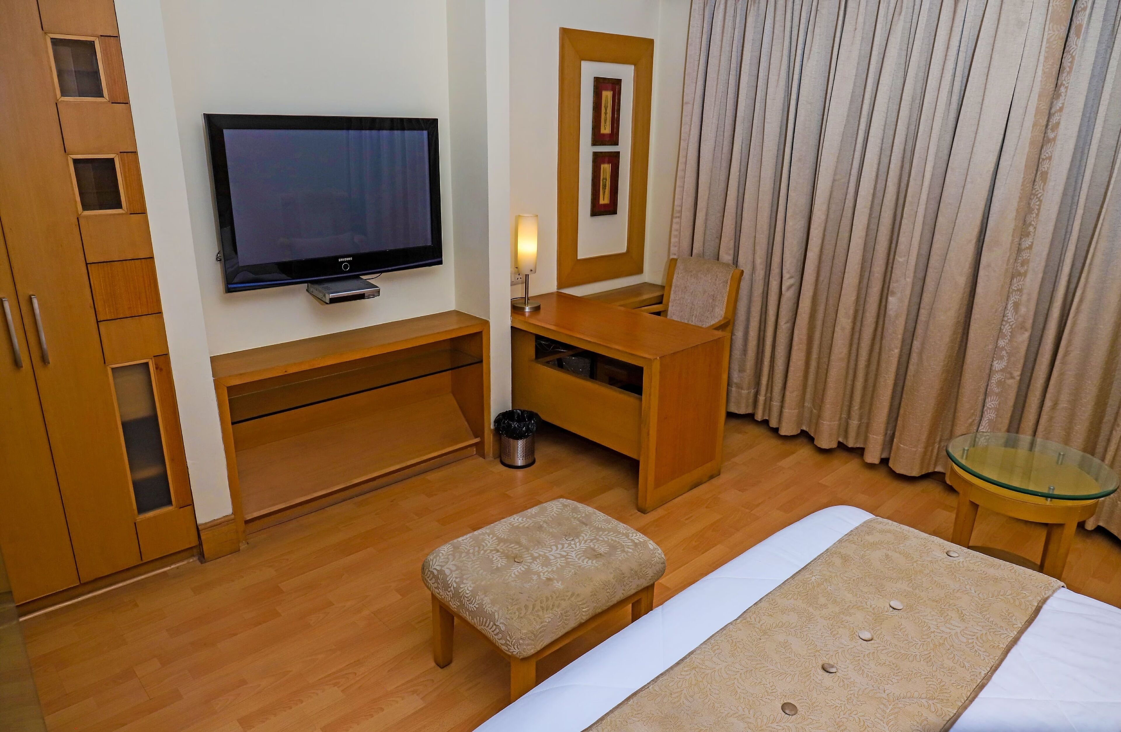 A hotel room with a television, wooden furniture, a glass table, a bench, and a bed.