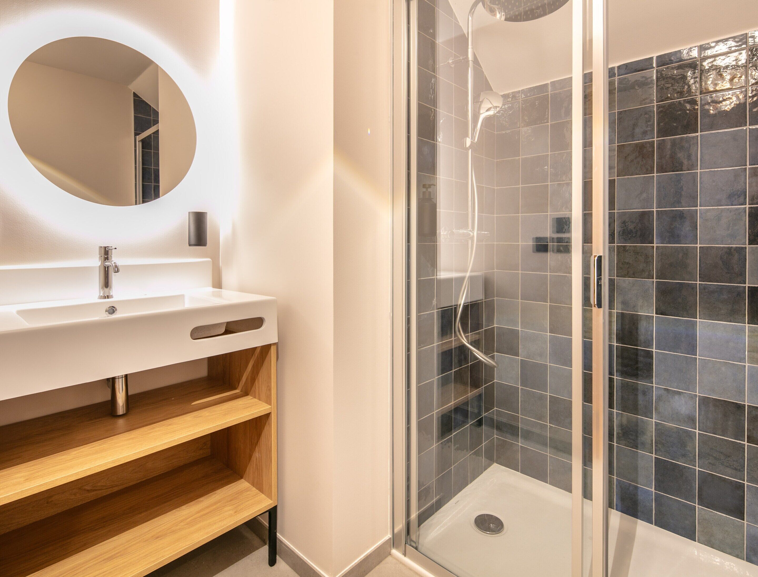 Une salle de bain moderne avec un lavabo blanc, des étagères en bois, une cabine de douche en verre et un miroir.