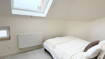 A bedroom with a skylight, a radiator, a bed with pillows, a bedside table with a vase, and a view of buildings outside.