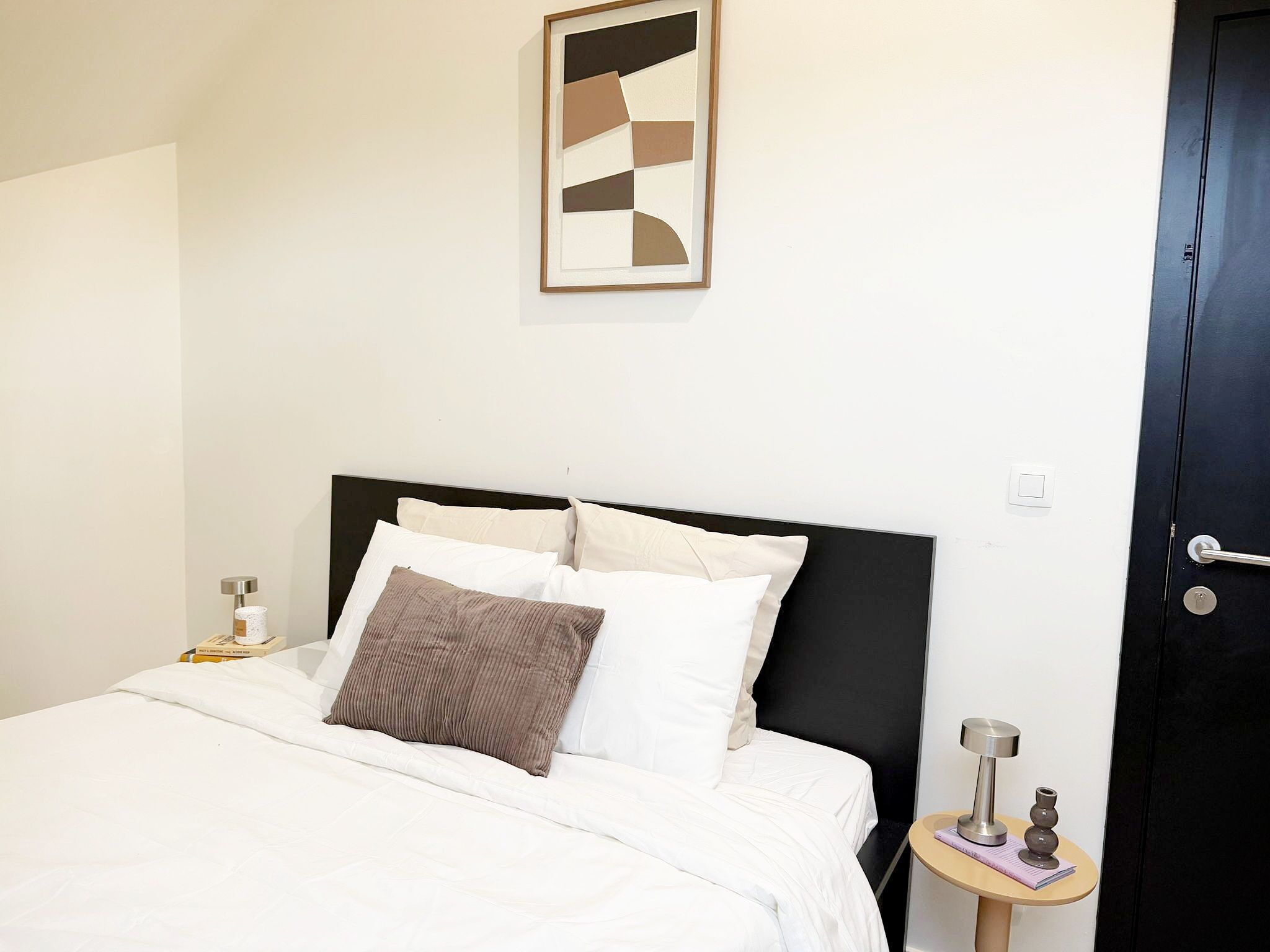 Una cama bien hecha con ropa de cama blanca y marrón, un cabecero negro moderno, una obra de arte abstracta enmarcada en la pared y una mesita de noche con una lámpara y objetos decorativos.