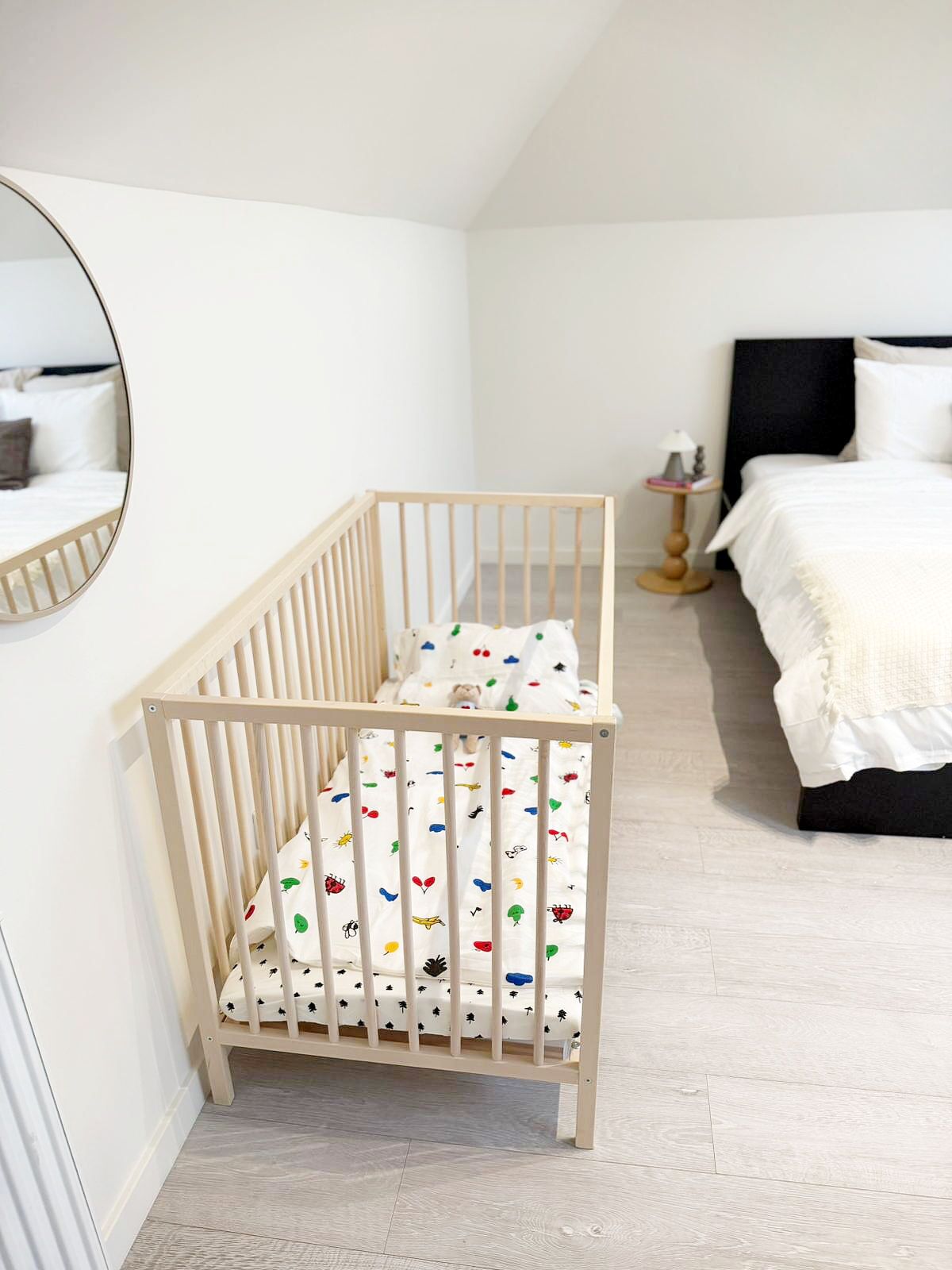 Un berceau blanc avec un matelas aux motifs colorés, une table de chevet avec une lampe, et un lit avec une literie blanche, dans une pièce aux murs et au sol blancs.