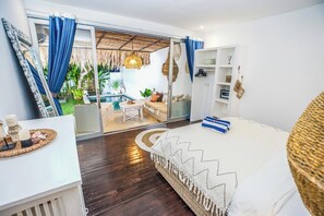 Ein Schlafzimmer mit einem großen Bett, einem kleinen Tisch, einem weißen Kleiderschrank und einer Glasdoor, die auf einen Balkon mit Pool und Sitzgelegenheit führt.