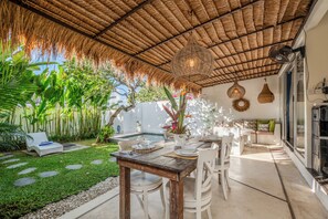 Un espace de restauration en plein air doté d’un toit de chaume, entouré d’une végétation luxuriante et d’une piscine.