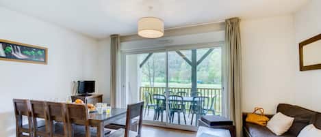 A room with a dining table, chairs, a sofa, a TV, and a large window with a view of greenery.