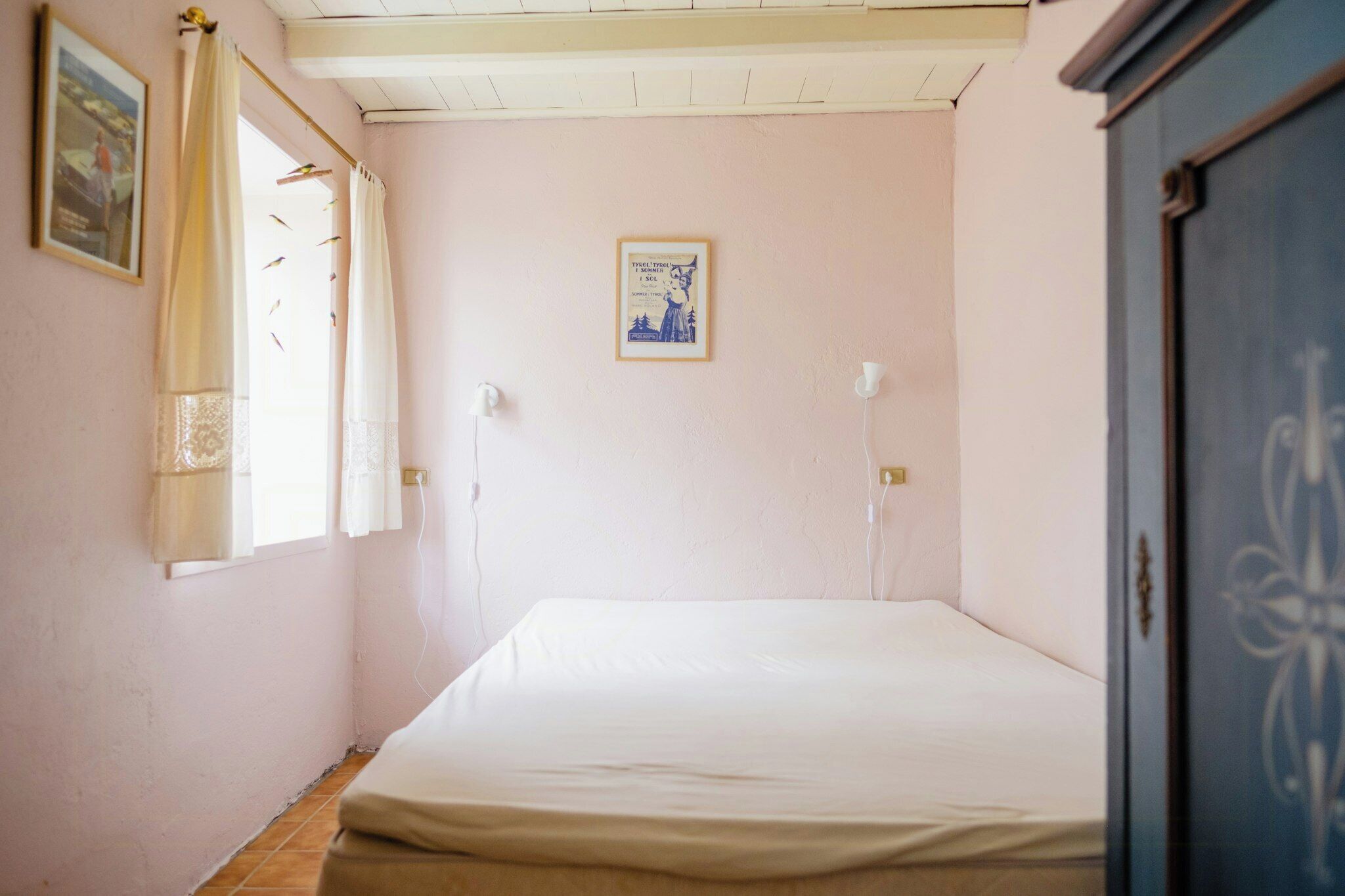 Une chambre avec un lit simple, une fenêtre avec des rideaux, un interrupteur fixé au mur et un tableau encadré.