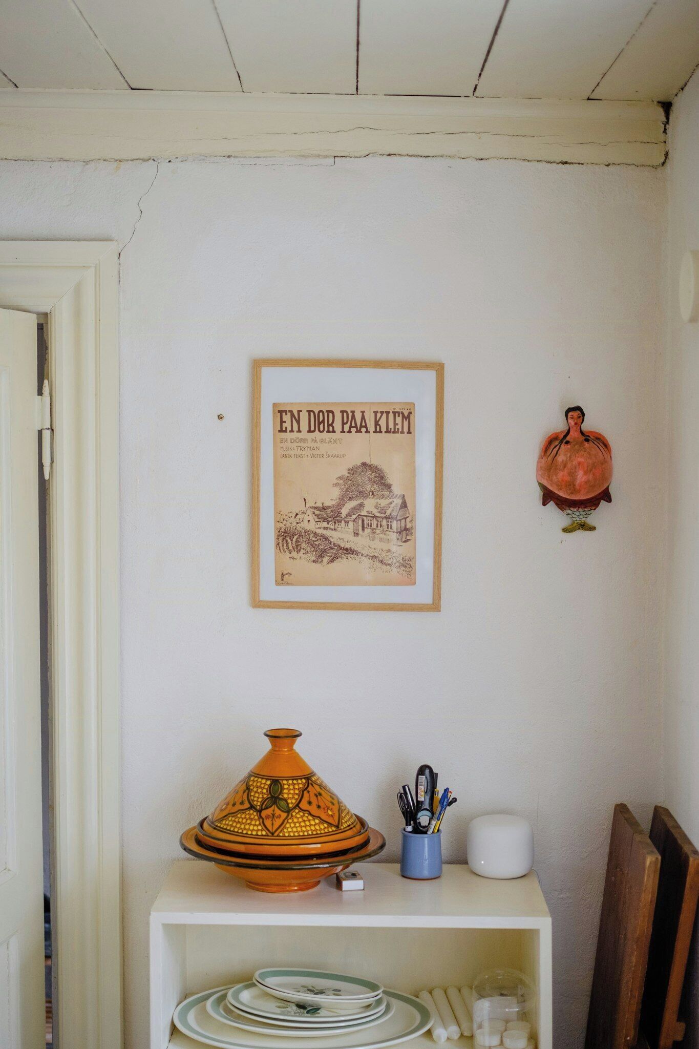 A room with a framed picture, a Moroccan tagine, and a shelf with plates and utensils.