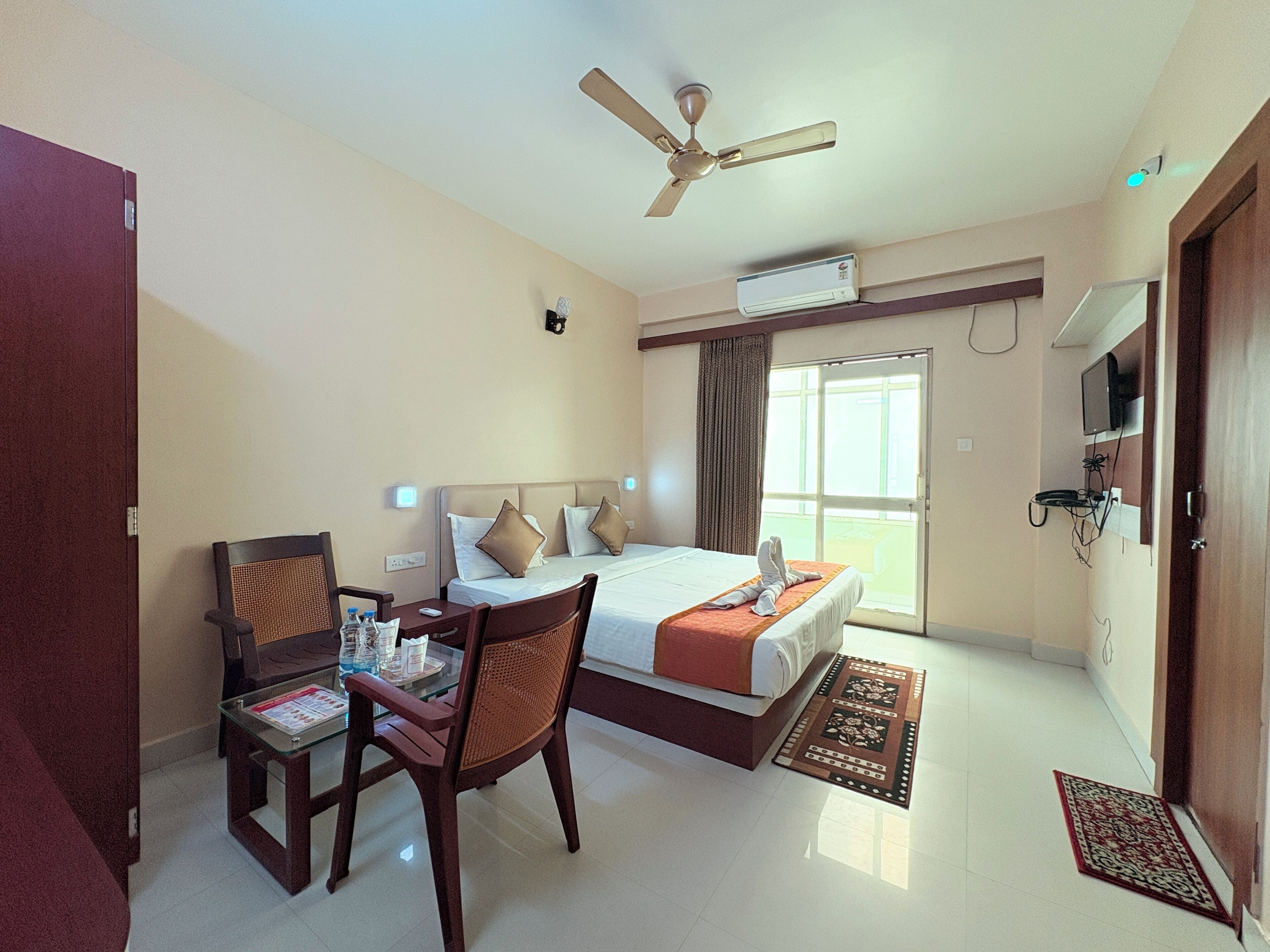 A hotel room with a bed, a desk with a chair, a ceiling fan, an air conditioning unit, a television, and a window with curtains.