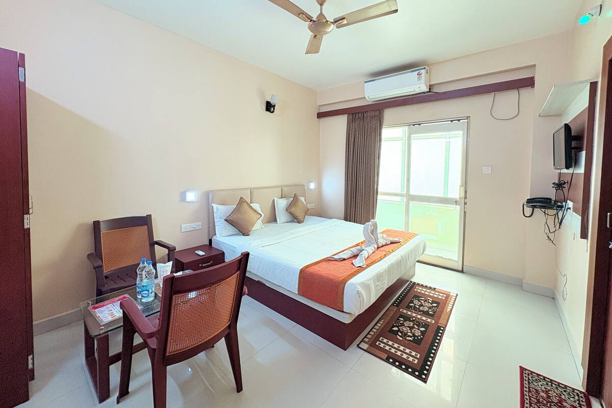 A hotel room with a bed, a desk with a chair, a ceiling fan, an air conditioning unit, a television, and a window with curtains.