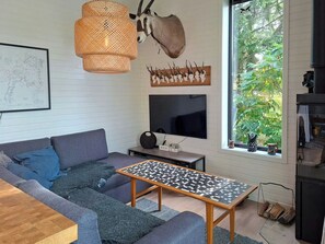 A modern living room with a sofa, a coffee table, a television, and a large window with a view of greenery. - Cottage with lake view and boat at Lake Storsjön (Sandviken)