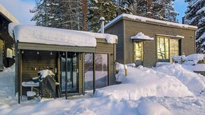 A modern cabin with a glass door entrance, a covered outdoor seating area, and a snow-covered roof.