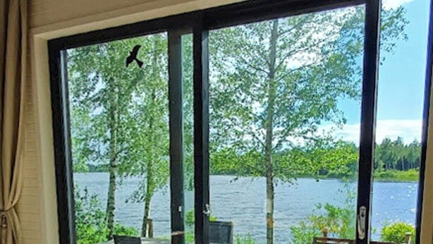 A room with a wooden table and chairs, a large sliding glass door leading to an outdoor deck with a view of a lake and trees. - Cottage with lake view and boat at Lake Storsjön (Sandviken)
