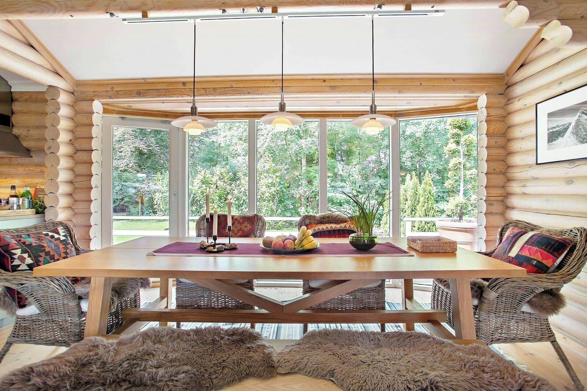 A log cabin interior with a wooden dining table, wicker chairs, a fur rug, and a large window with a view of greenery.