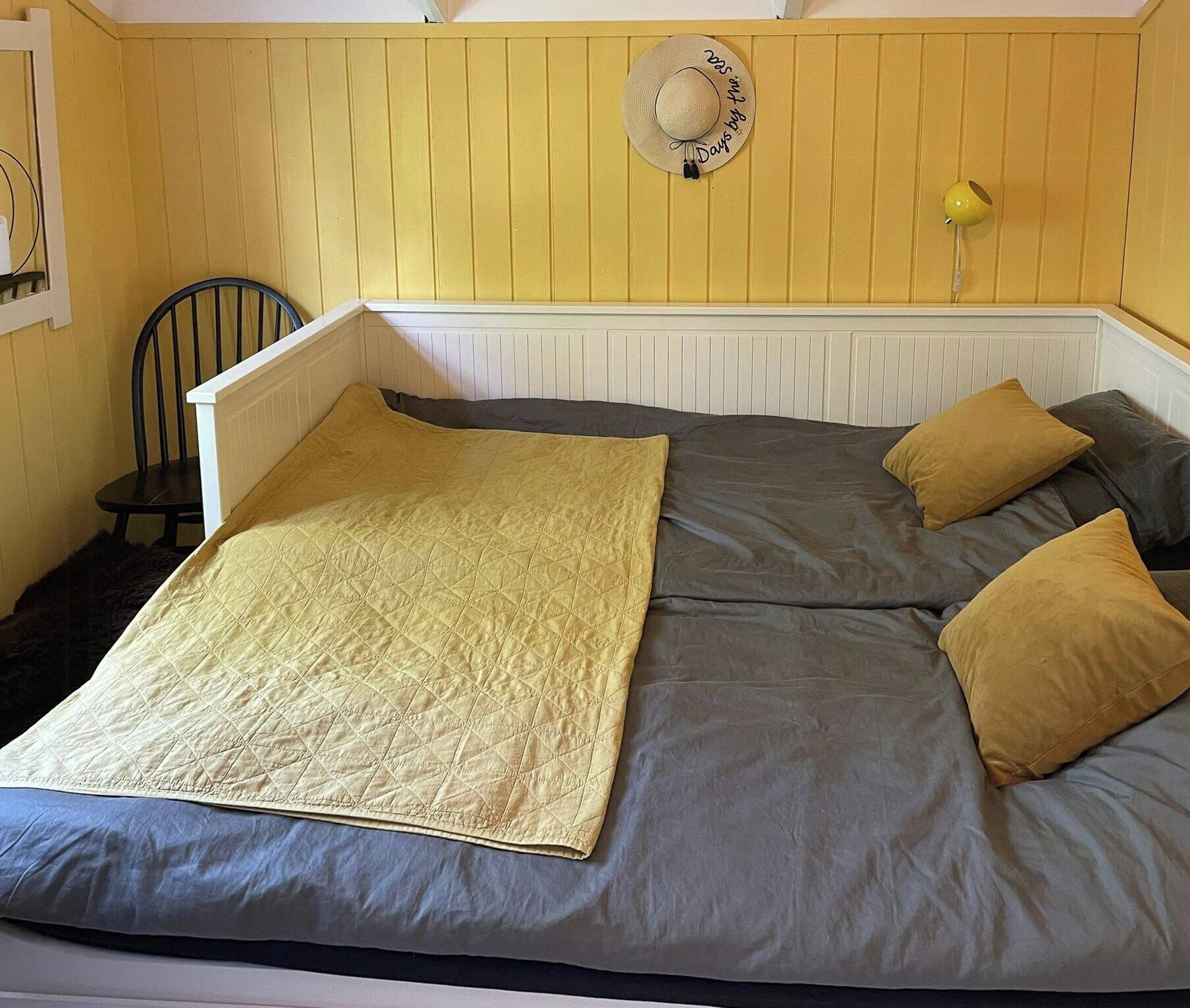 A bed with a quilt, two pillows, a chair, a hat on the wall, and a lamp.