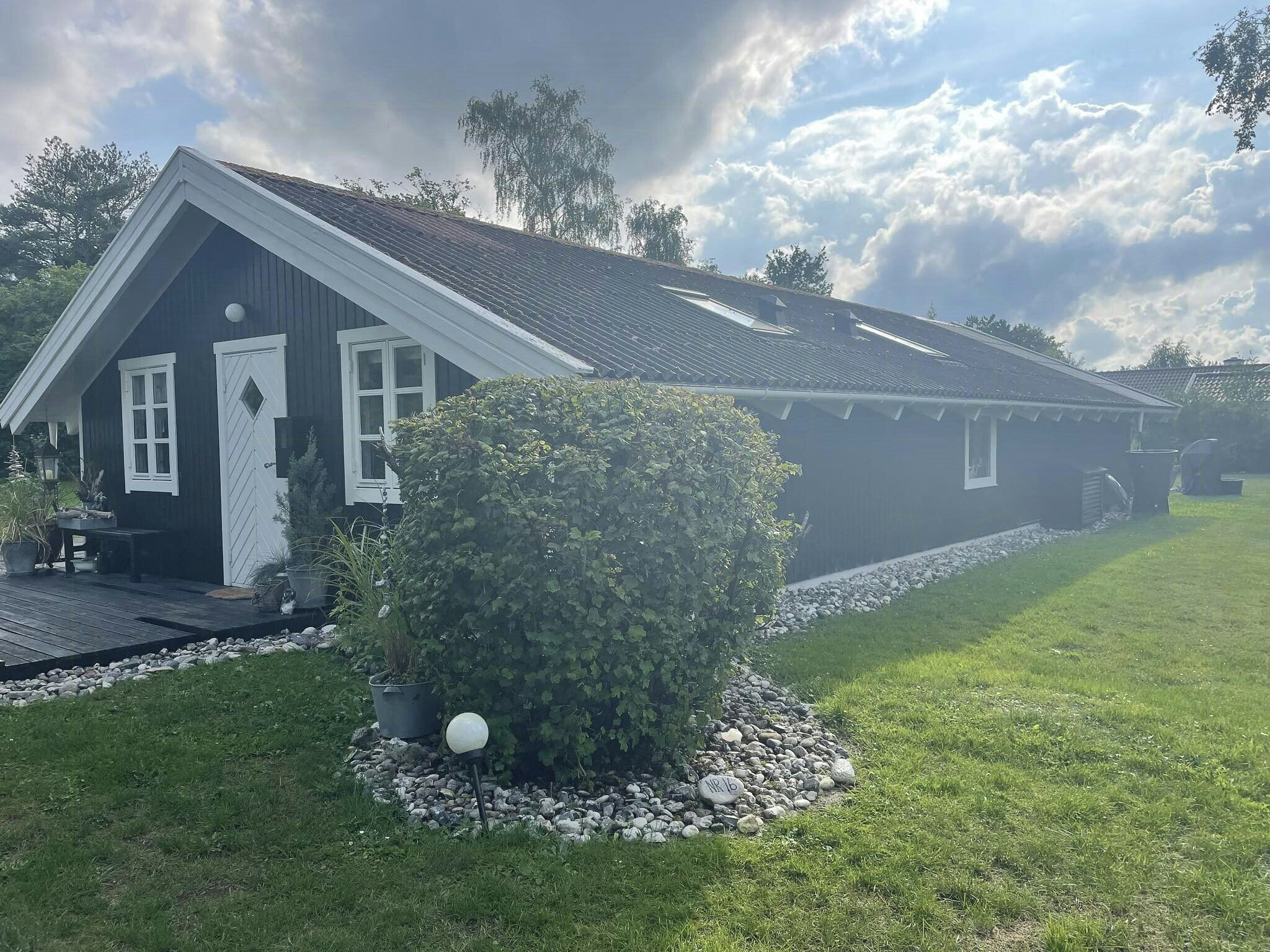 Une maison aux couleurs sombres, avec un toit en pente, un porche à l’avant et un petit jardin agrémenté d’un arbuste et d’un lampadaire.