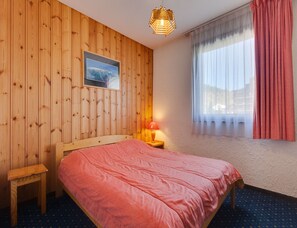 A wooden-paneled room with a bed, a nightstand, a lamp, a framed picture, and a window with curtains. - Slope-side Apartment With Balcony Views (Les Deux Alpes)