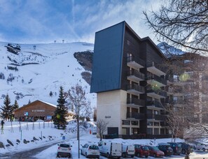 A modern building with balconies, a ski slope in the background, and a parking area with cars. - Slope-side Apartment With Balcony Views (Les Deux Alpes)