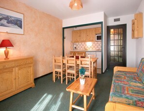 A cozy living room with a wooden table, chairs, a sofa, a lamp, and a kitchen in the background. - Slope-side Apartment With Balcony Views (Les Deux Alpes)