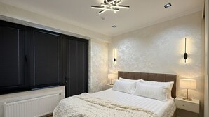A bedroom with a bed, bedside tables, wall sconces, a radiator, and a window with blinds. - Modern Apartment in Alfraganus  Rent Car & Moto (Tashkent)