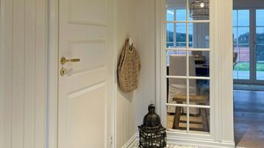 Un couloir avec une porte blanche, un sac tressé suspendu, une lanterne et, à travers des portes vitrées, une vue sur un espace extérieur.