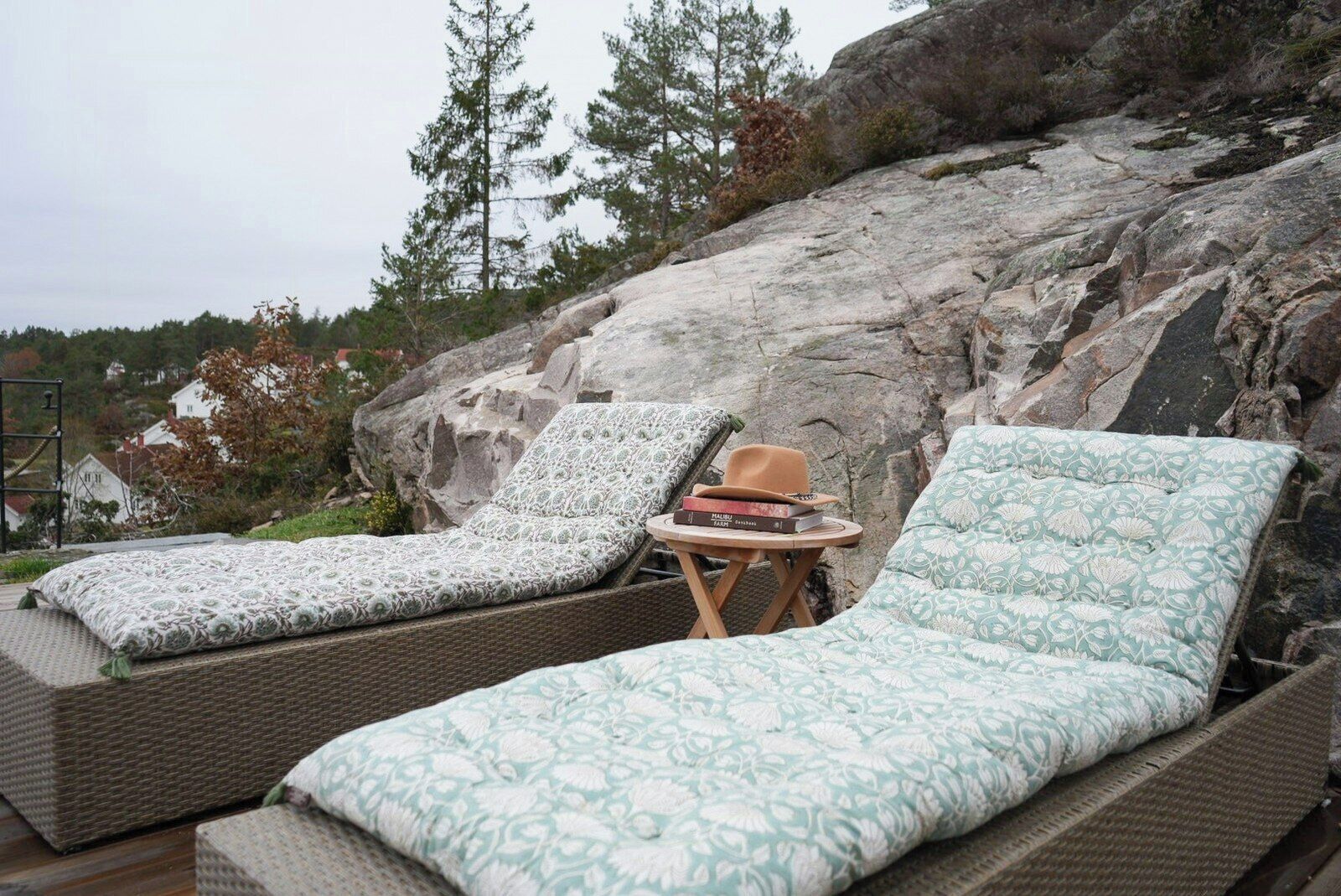 Two wicker lounge chairs with patterned cushions, a small wooden table with a hat and books, and a rocky backdrop with trees.