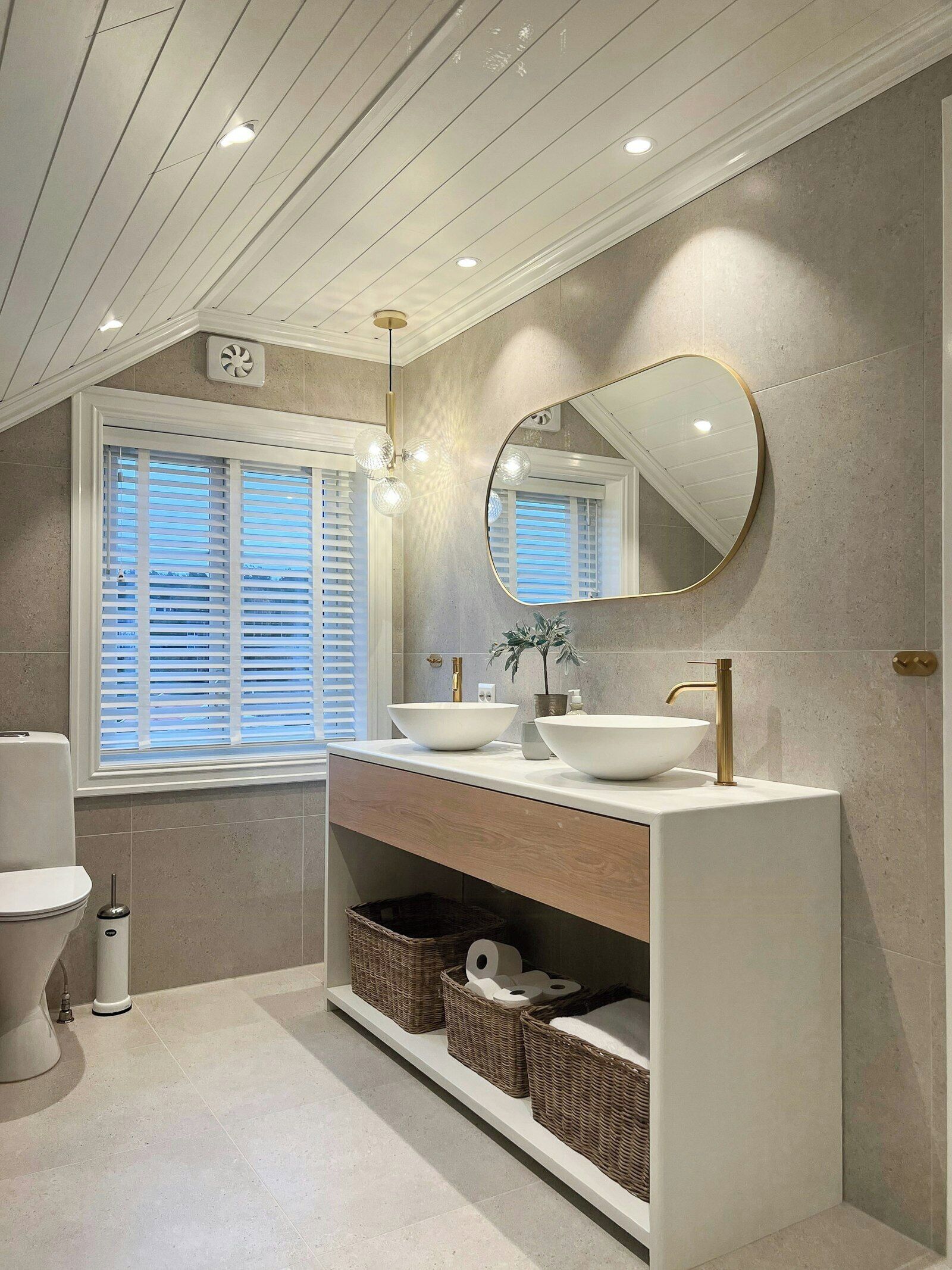 A modern bathroom with a white vanity, two sinks, a round mirror, and a window with blinds.