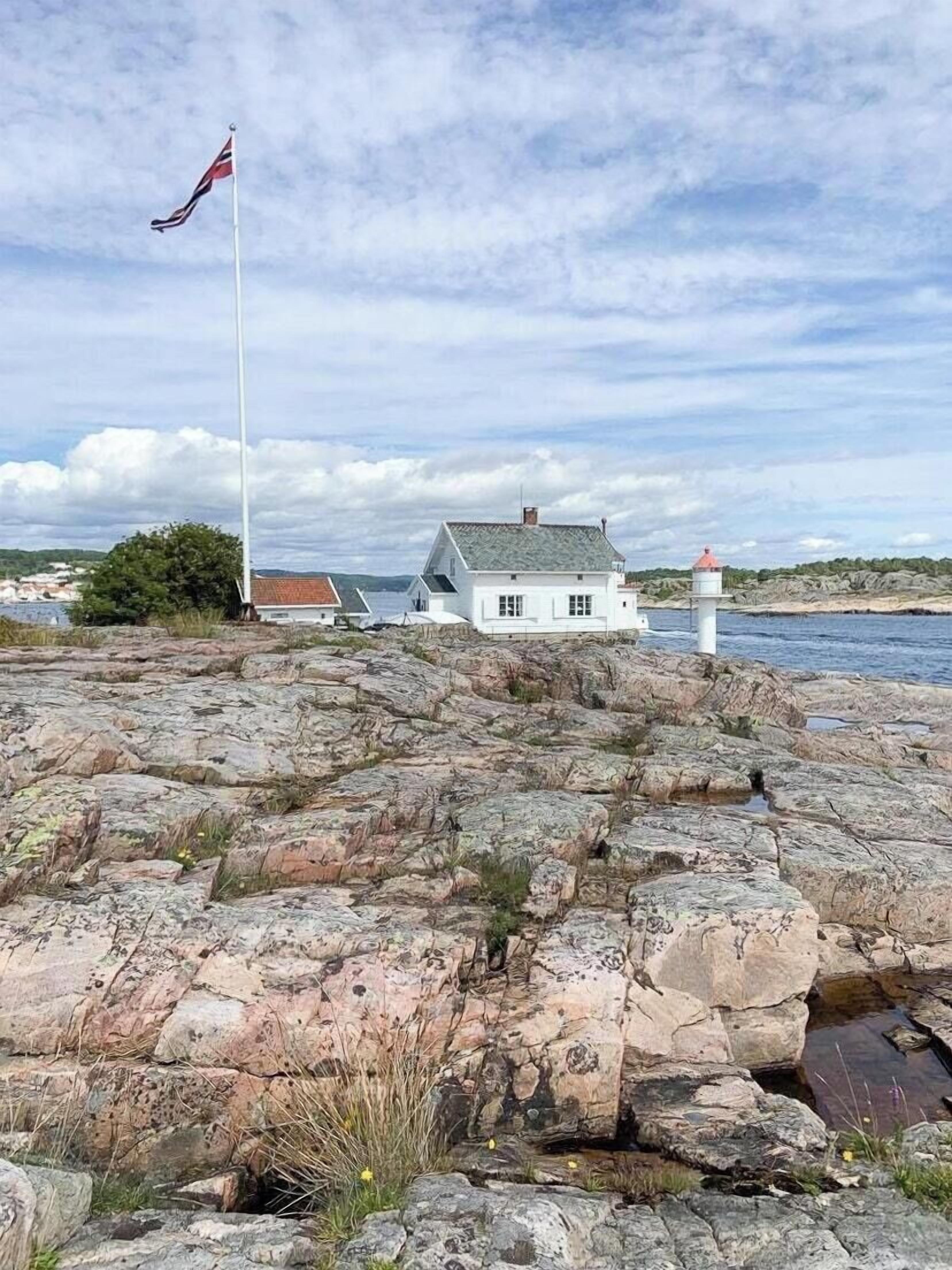 Una casa costera con un faro al fondo, terreno rocoso e una bandera en un asta.