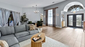 A modern living room with a grey sofa, a wooden coffee table, and a chandelier. - Screened Pool + Firepit + Outdoor Lounge | Mins to WEF + Polo | Palm & Pool (Wellington)