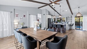 A spacious dining area with a wooden table and modern chairs, a kitchen with a bar, and a view of a pool. - Screened Pool + Firepit + Outdoor Lounge | Mins to WEF + Polo | Palm & Pool (Wellington)