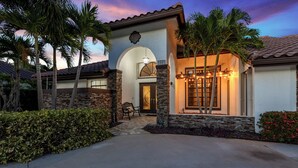 A house with a stone facade, a covered porch, and a well-lit entrance. - Screened Pool + Firepit + Outdoor Lounge | Mins to WEF + Polo | Palm & Pool (Wellington)