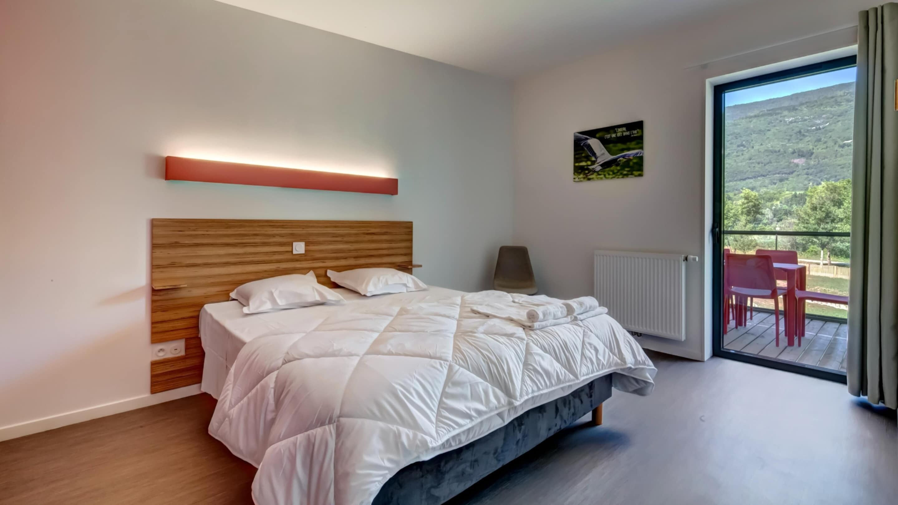 A modern bedroom with a large bed, a bedside table, a chair, a radiator, and a balcony with a view.