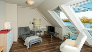 Et moderne loftværelse med skråvægge, stort ovenlys, en grå sofa, en hvid lænestol, et lille bord med en vinflaske og et fjernsyn på et stativ.