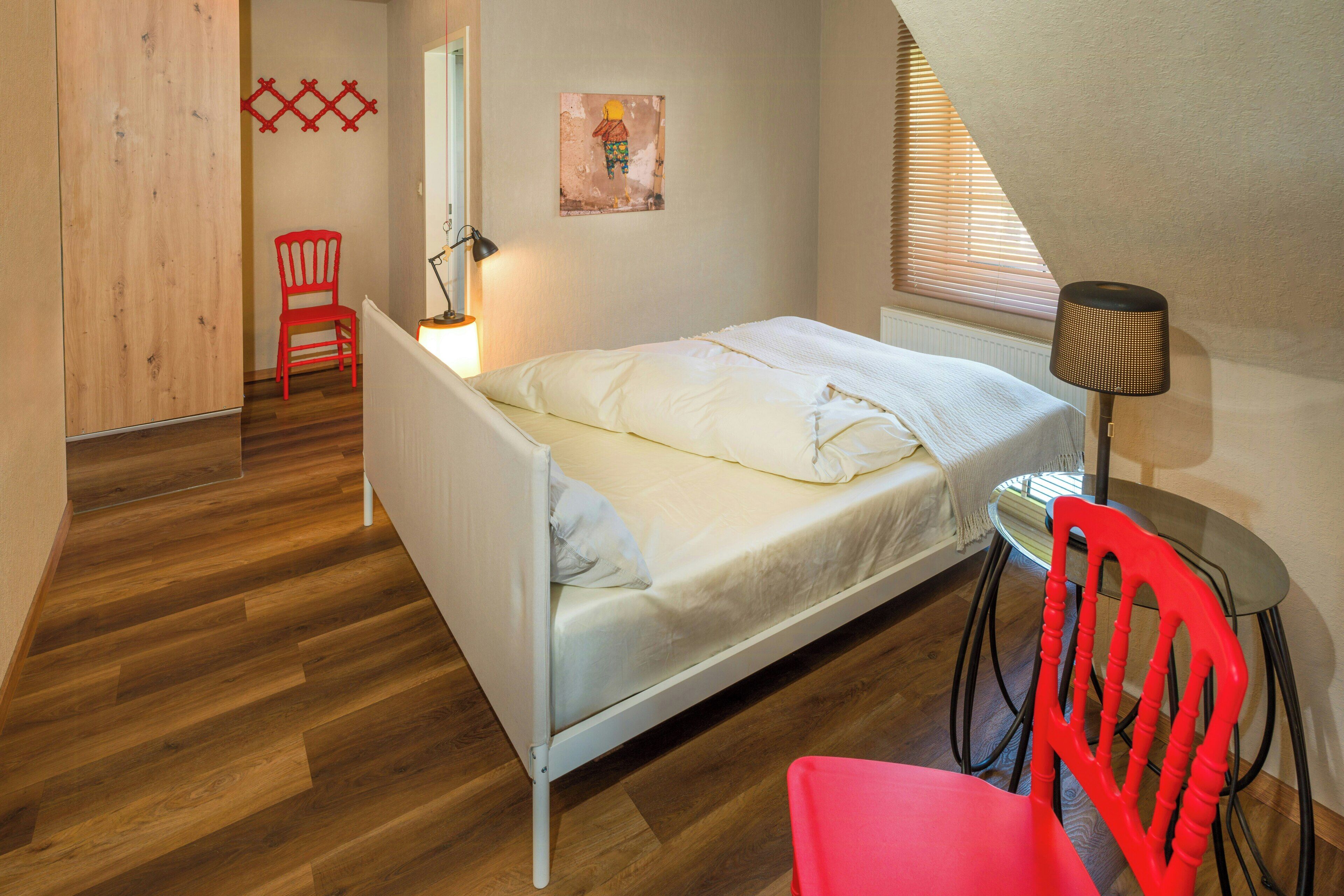 Una camera da letto con un letto, una sedia rossa, una lampada e un quadro appeso al muro.