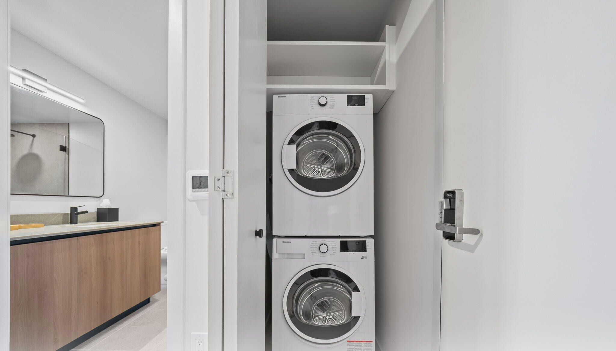 A modern laundry room with two stacked front-loading washing machines, a wooden vanity with a mirror, and a light fixture above the mirror.