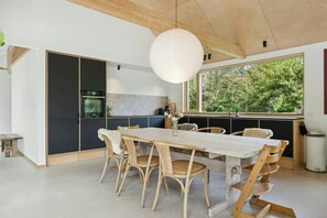 A modern kitchen with a dining area, featuring a large window with a view of greenery, a hanging light fixture, and a dining table with chairs.
