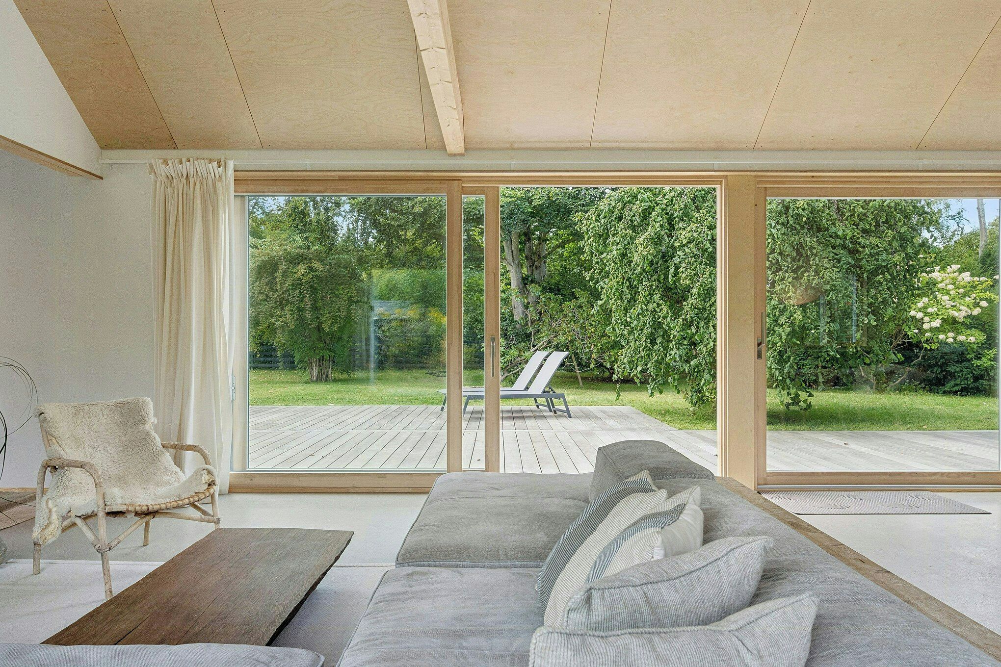 A modern living room with a large sofa, a wooden coffee table, and a wicker chair. There are large sliding glass doors leading to an outdoor deck.