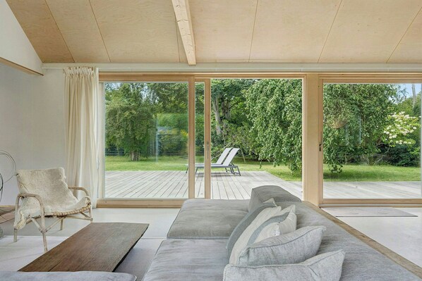 A modern living room with a large sofa, a wooden coffee table, and a wicker chair. There are large sliding glass doors leading to an outdoor deck.