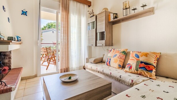A living room with a sofa, a coffee table, a bookshelf, and a sliding glass door leading to an outdoor area. - Chrysanthos Maisonette by RentalsPro - Siviri Halkidiki (Siviri)