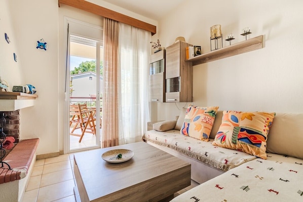 A living room with a sofa, a coffee table, a bookshelf, and a sliding glass door leading to an outdoor area. - Chrysanthos Maisonette by RentalsPro - Siviri Halkidiki (Siviri)