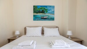 A neatly made bed with white linens, two pillows, and two folded towels. There are two bedside tables with lamps. A painting of a tropical beach scene hangs above the bed. - Chrysanthos Maisonette by RentalsPro - Siviri Halkidiki (Siviri)