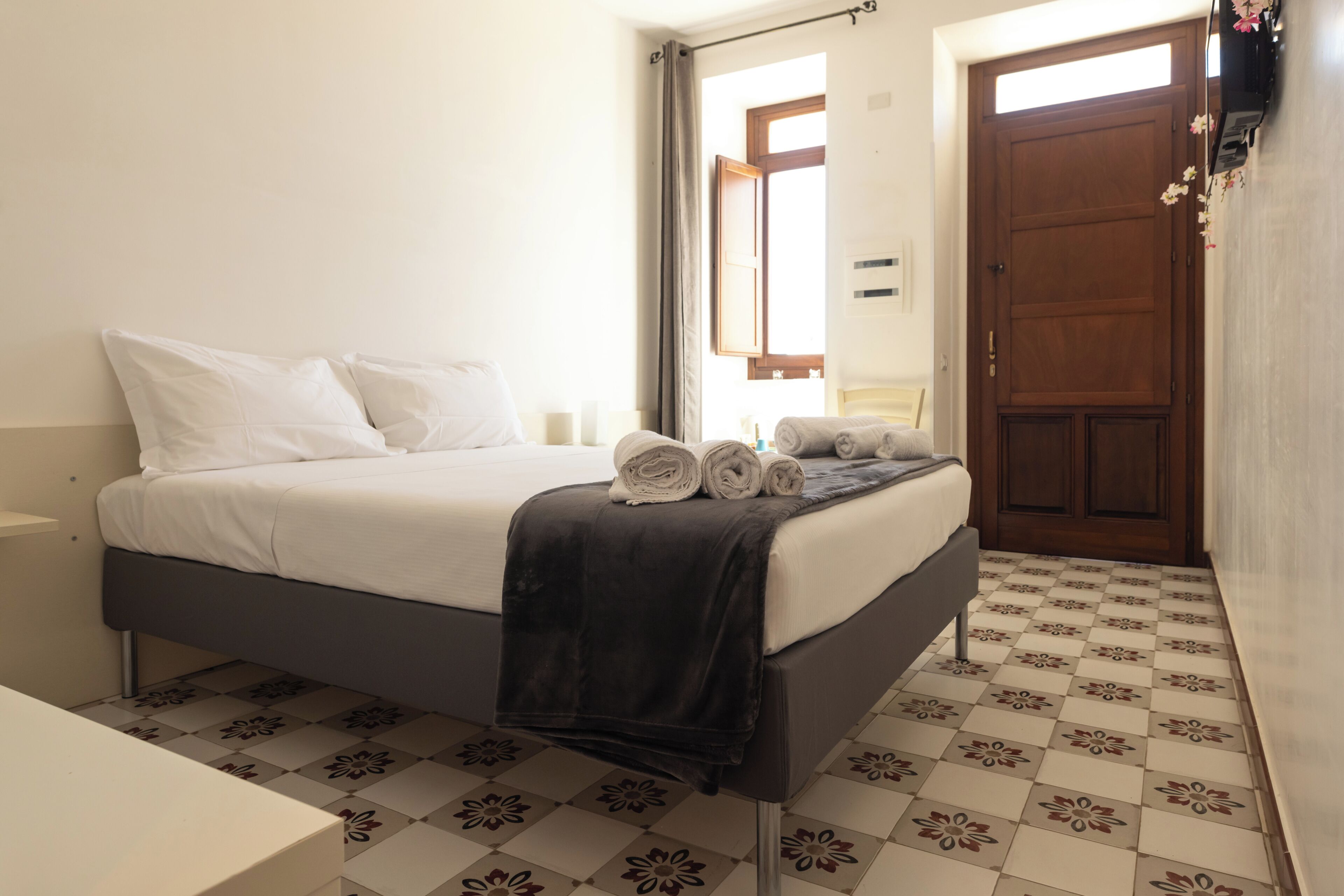 Un dormitorio con cama, mesita de noche, ventana con cortinas, puerta y suelo de baldosas estampadas.